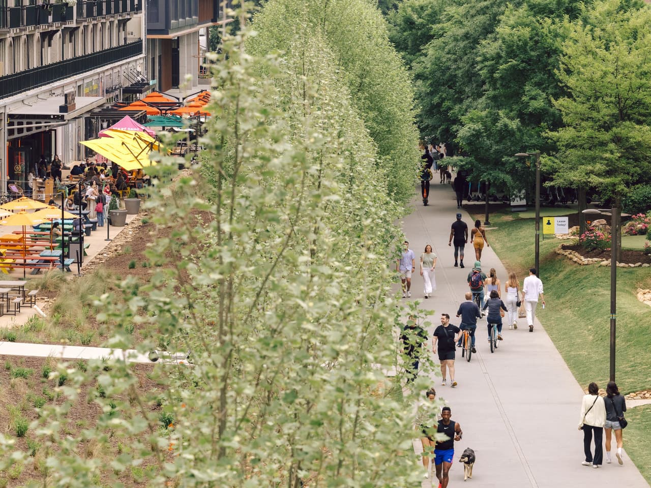 A crowd walks along a paved pathway. To their left are several restaurants and businesses.