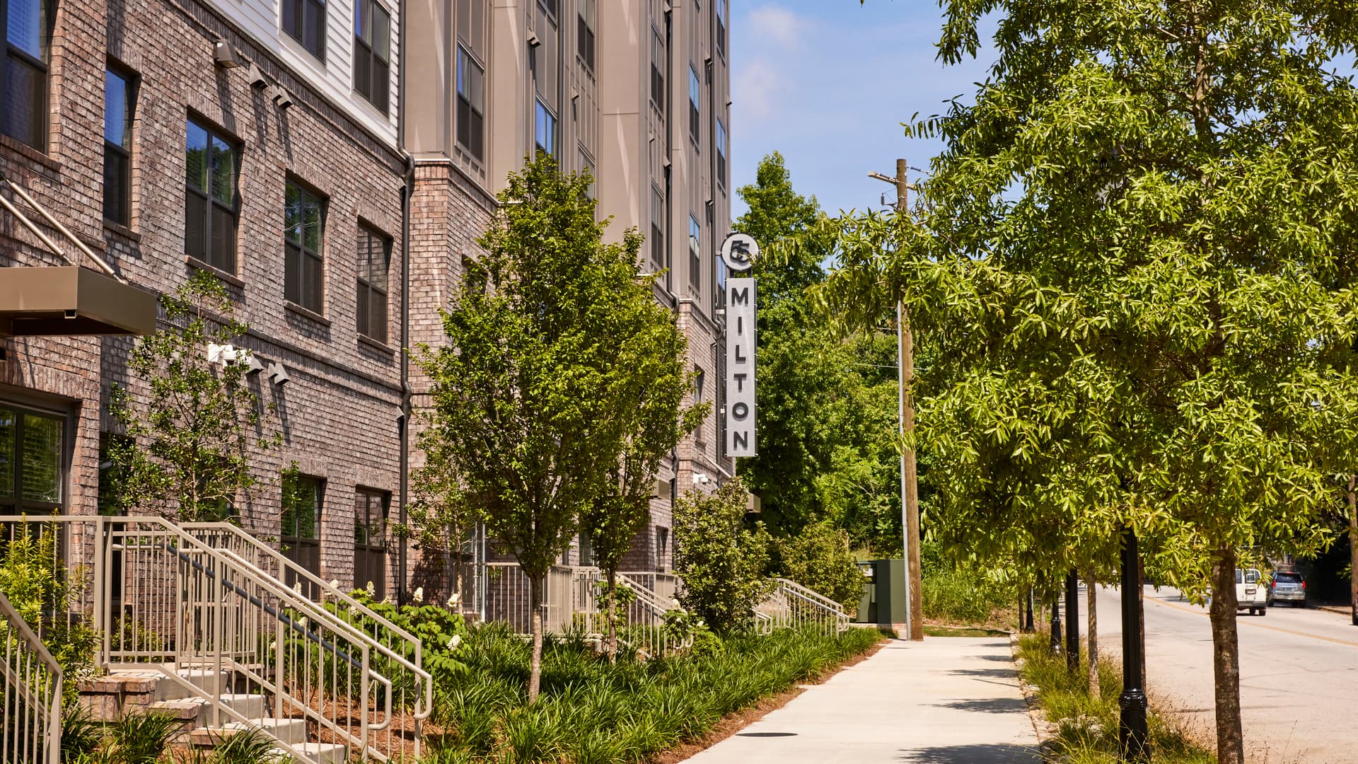 A brick apartment building sits in front of a sidewalk lined with green trees.