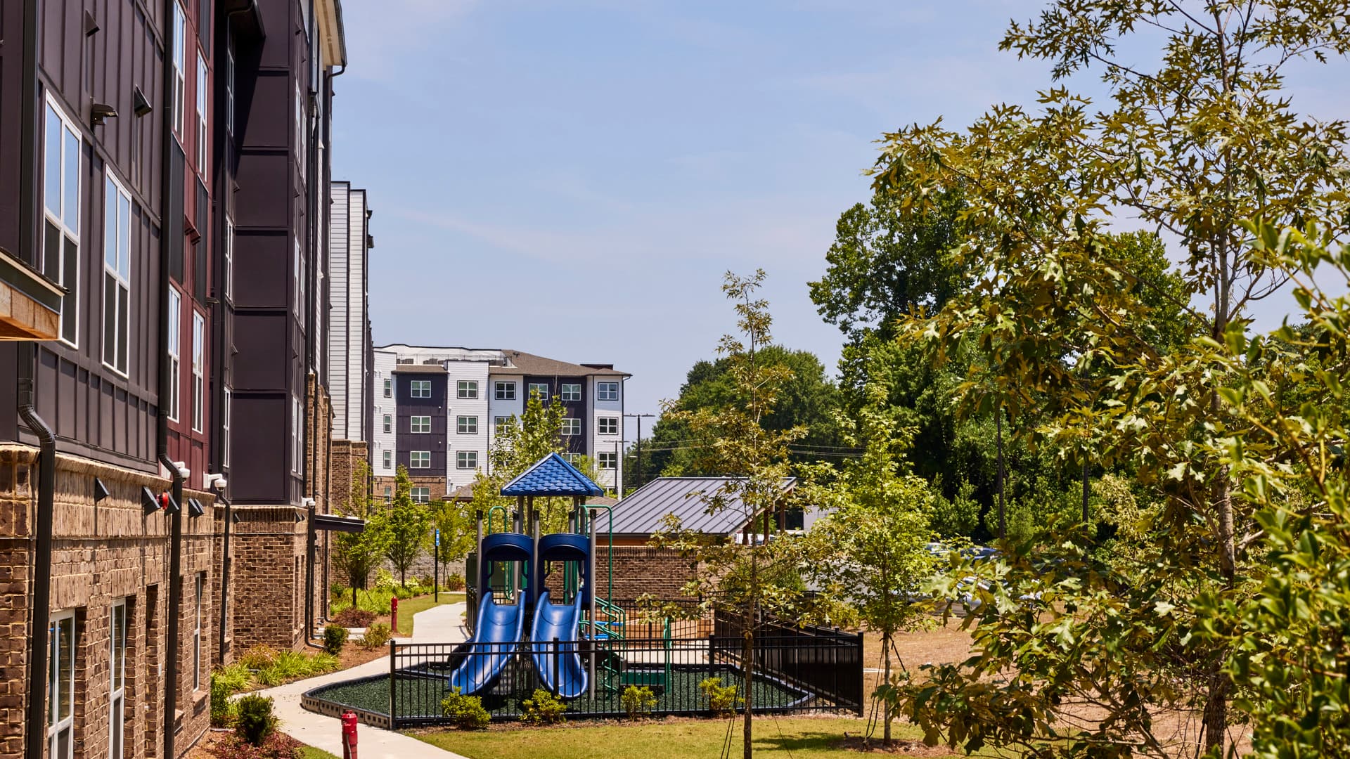 The back of an apartment building sits to the left of a nice sidewalk, a small playground, and a lot of greenery.