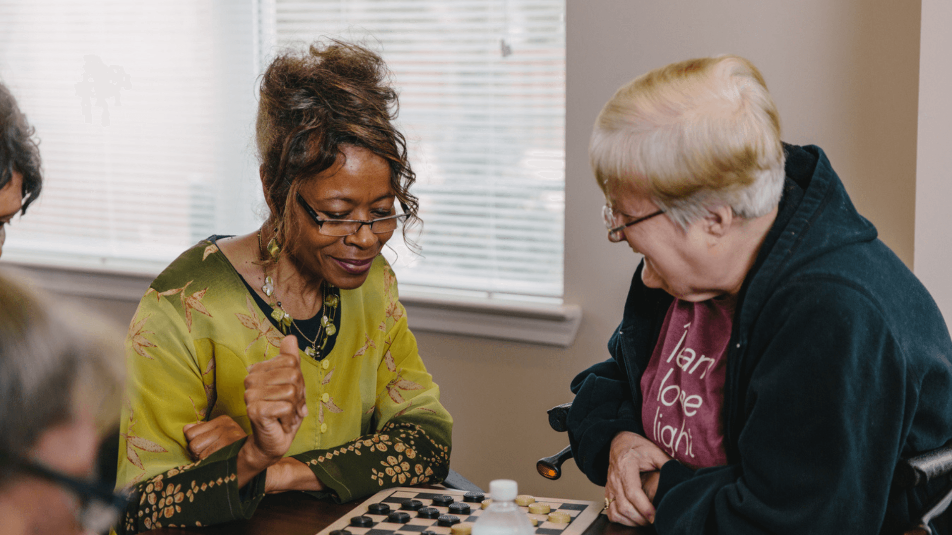 Two older women sit at a table playing checkers with each other.