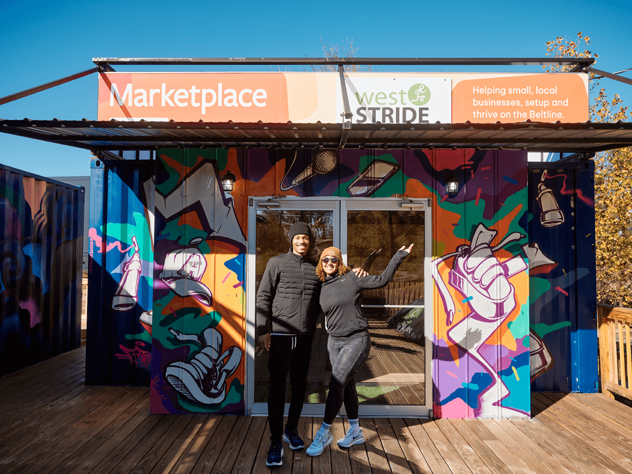 An African American couple stand proudly in front of a container storefront, showing off their space. The sign says Marketplace West Stride.